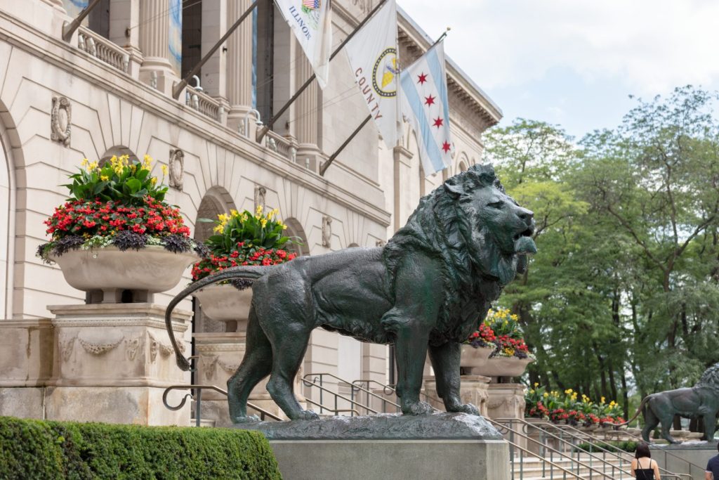 A lion sculpture standing near urns of flowers outside an imposing building with flags adorning it