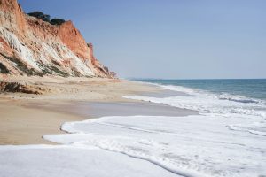 View of waves hitting a golden beach bordered by redstone cliffs, with a blue sky in the background.