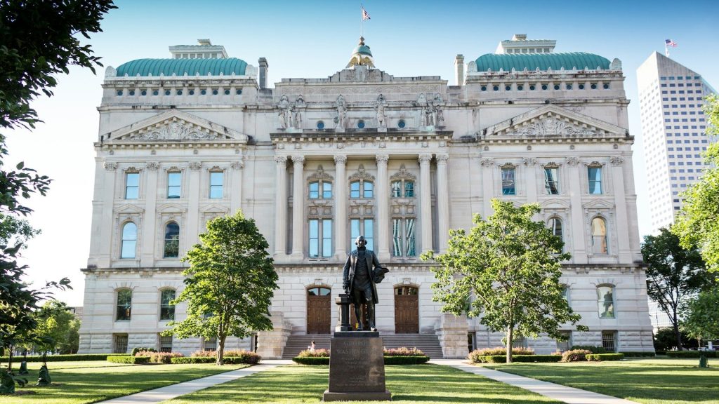 A historic statehouse building in Indianapolis with green leafy trees, lawn and a statue to the front and blue sky in the background.
