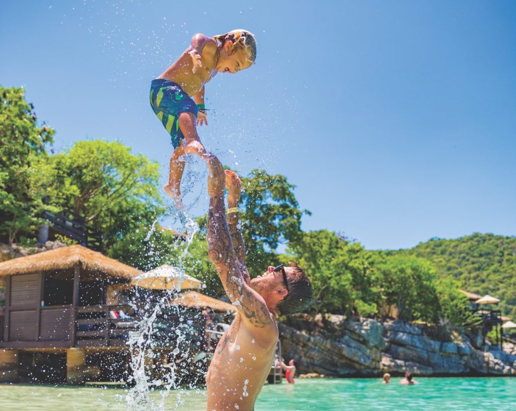 Father throwing his son into the air from the blue waters of a beach, blue sky in background