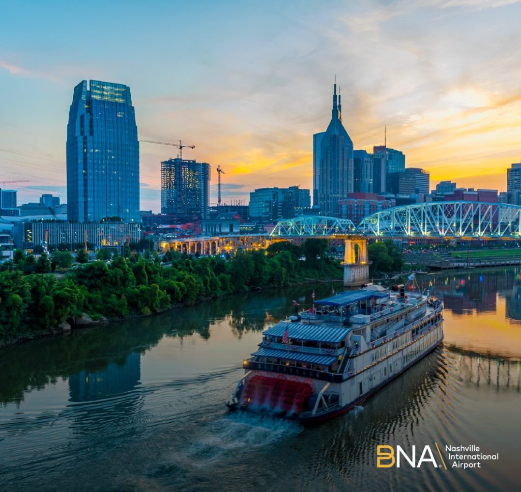 Ferry cruiser in the foreground on a river, with a city skyline, greenery and a sunset sky in the background.