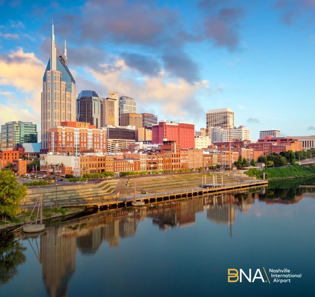 A river in the foreground, sunlit city skyline and blue sky in the background