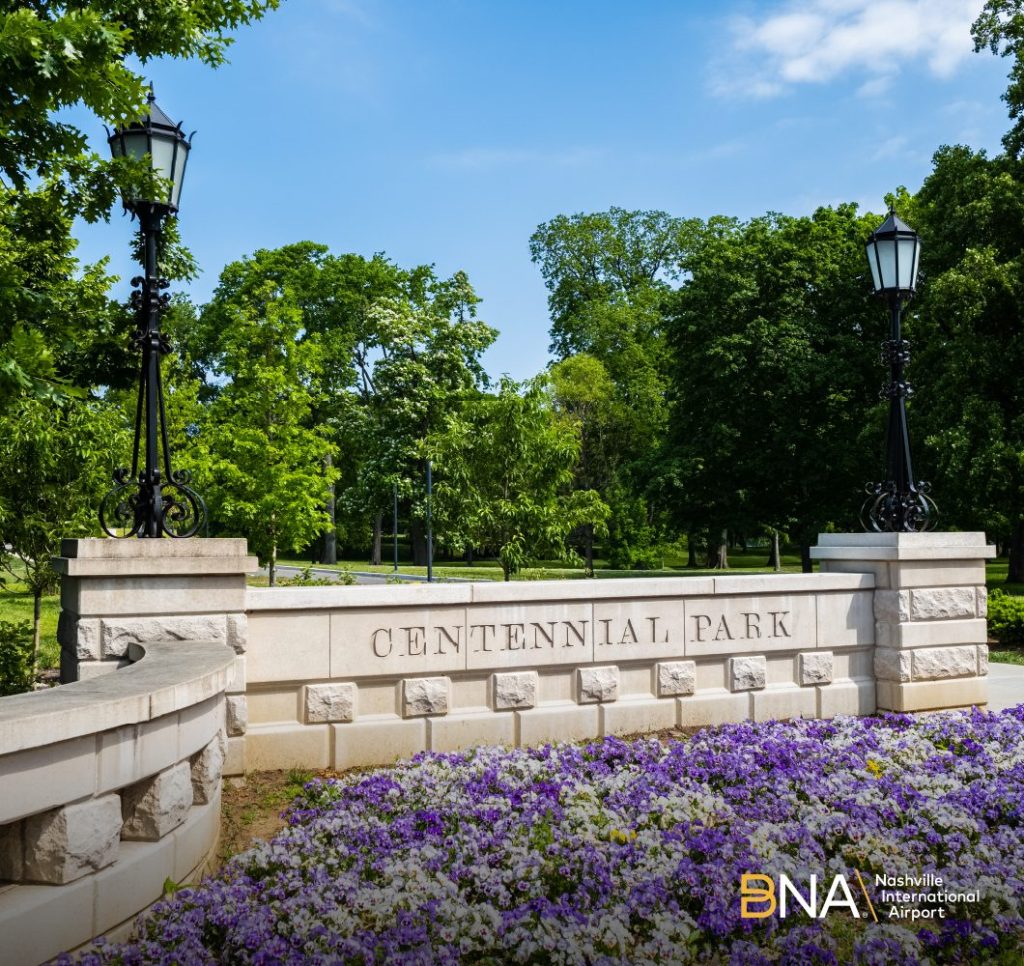 A wall bearing the inscription "centennial park" with flowers in the foreground and green trees in the background