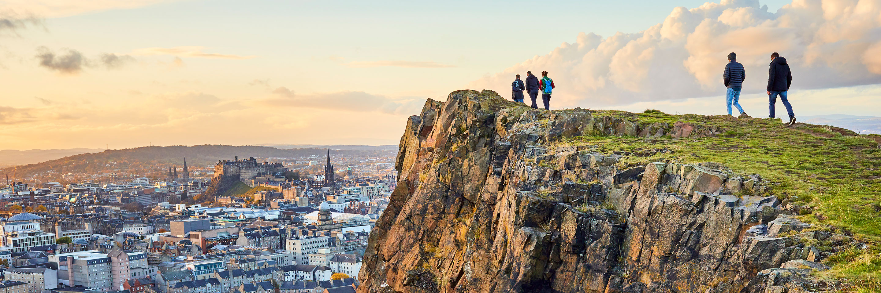Group of people walking along cliff edge at Salisbury Crags, Holyrood Park with Edinburgh city the in background at sunset